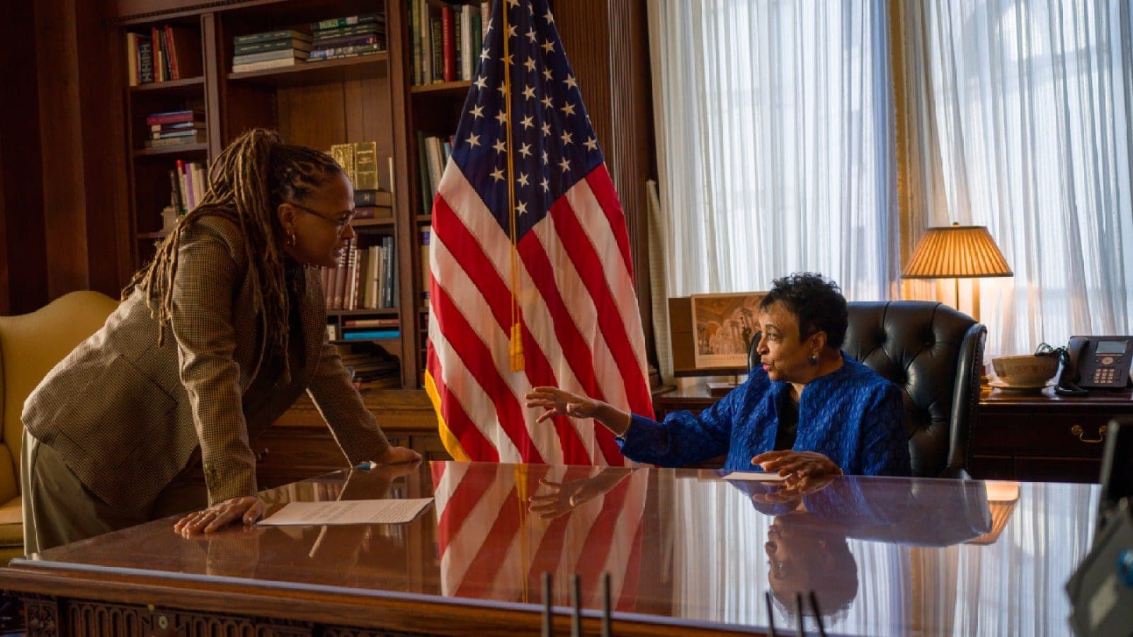 Ava DuVernay leans on a desk talking to Carla Hayden, who is seated beside a United States flag, in a still from 14th
