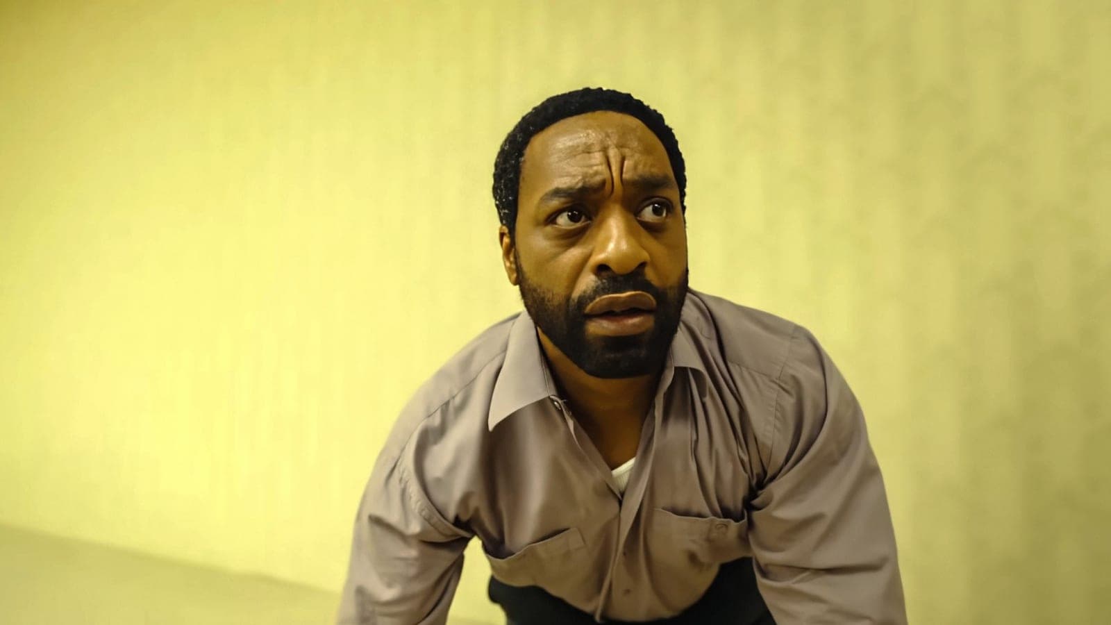 Chiwetel Ejiofor crouches against the yellow patterned wallpaper of an empty room, looking up in alarm, in a still from Backrooms.