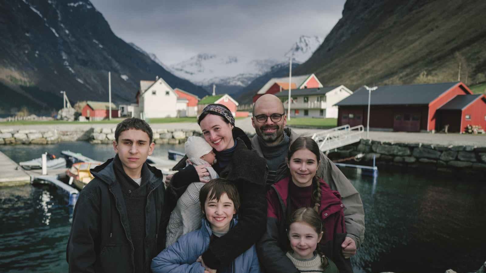 A family of seven pose together on a jetty in a Norwegian fjord village, red boathouses and snow-capped mountains behind them, in a still from Fjord.