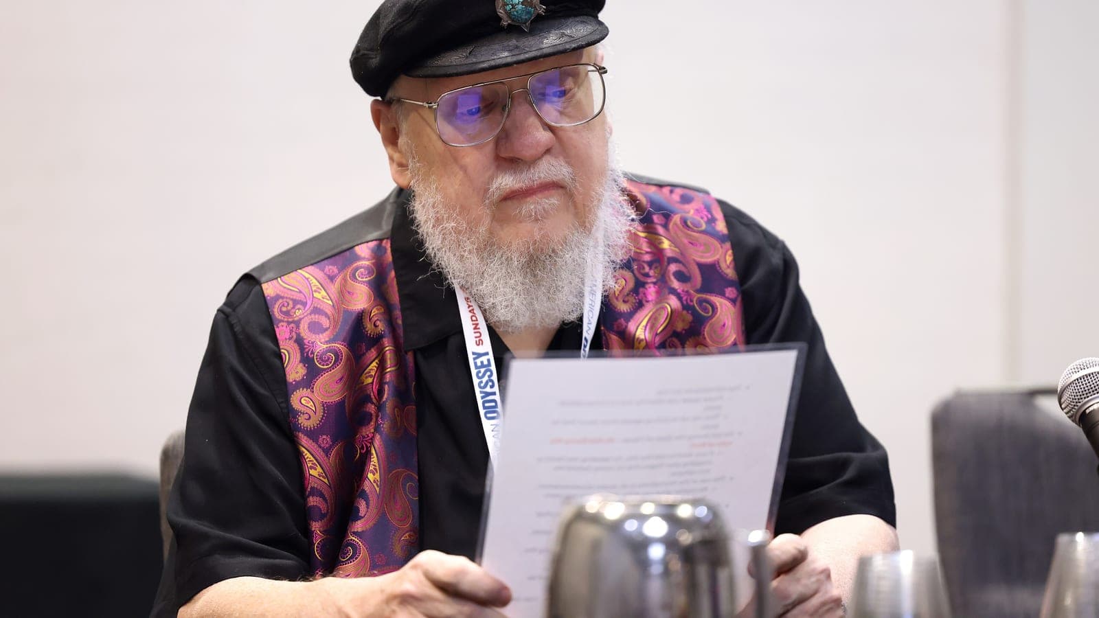 George R. R. Martin in a black cap and paisley waistcoat, reading from a printed page at a convention panel table
