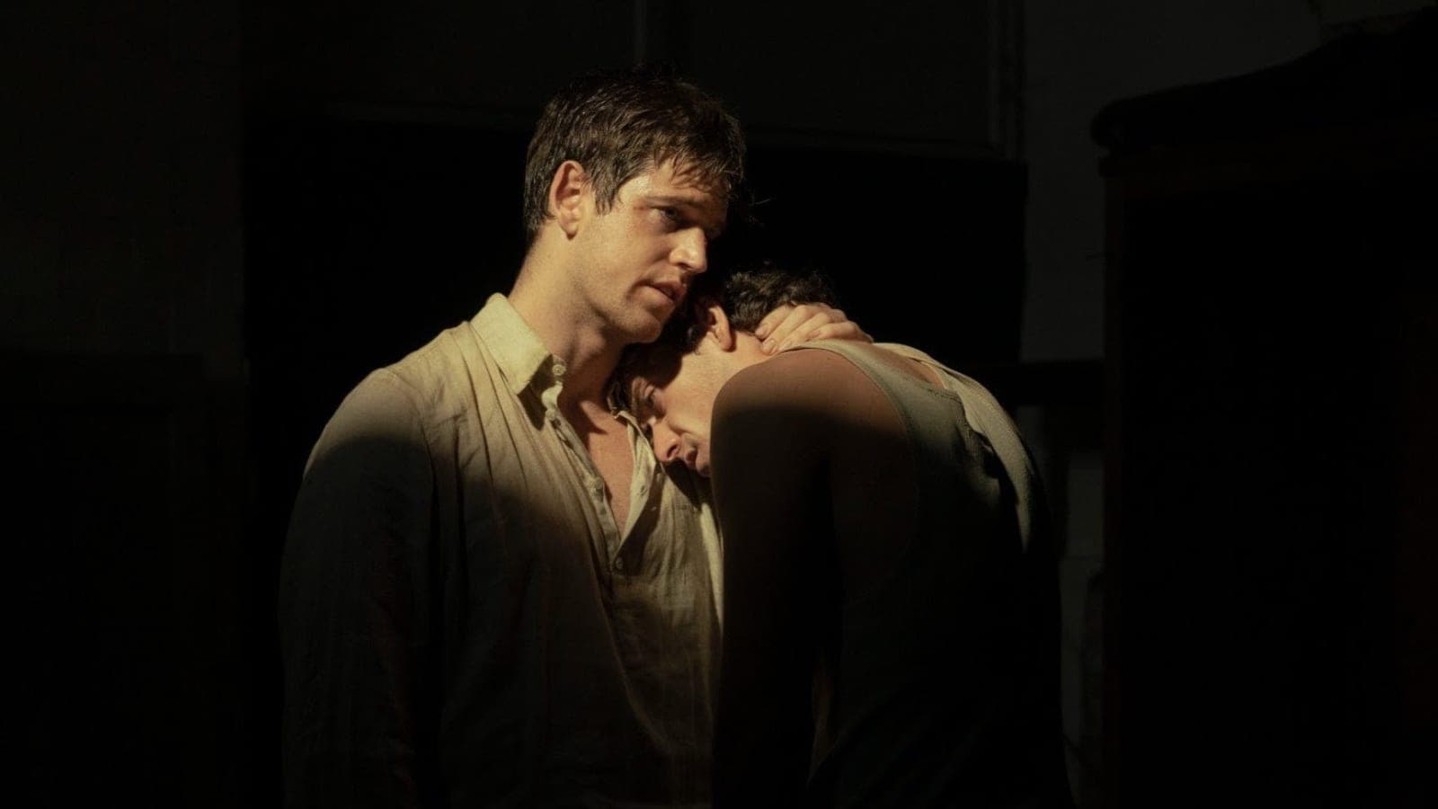 Two men stand close together in a dimly lit room, one in a pale collared shirt with his arm around the other, who rests his head against his chest, in a still from La Bola Negra.