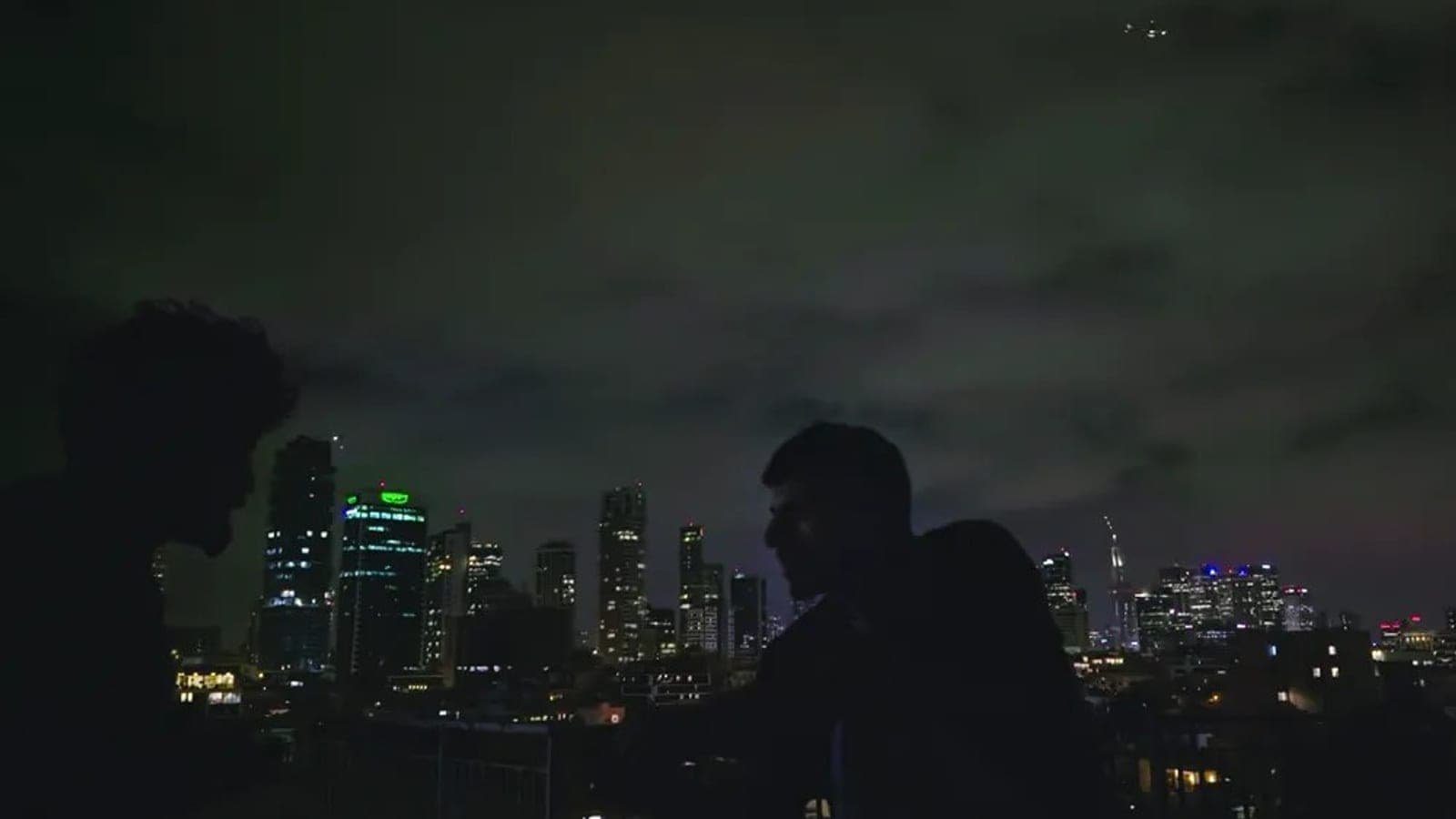 Two figures in silhouette on a rooftop at night, a lit city skyline behind them, in a still from the documentary NAZA