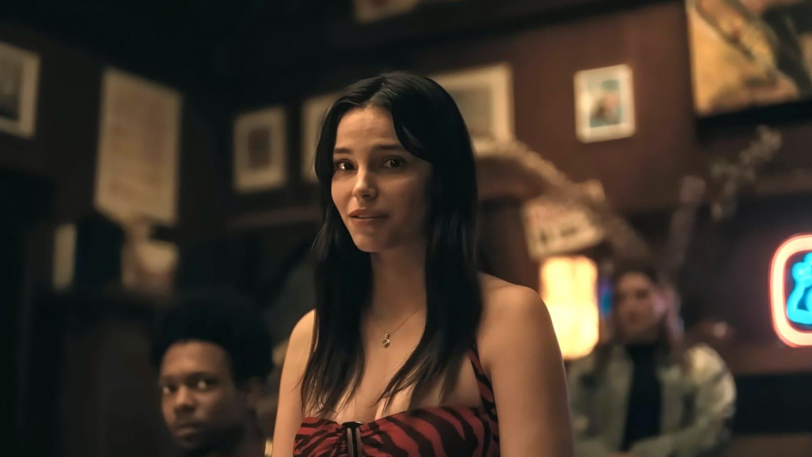Inde Navarrette in Obsession, standing in a dimly lit bar in a red striped top, with framed pictures and a neon sign on the wall behind her.