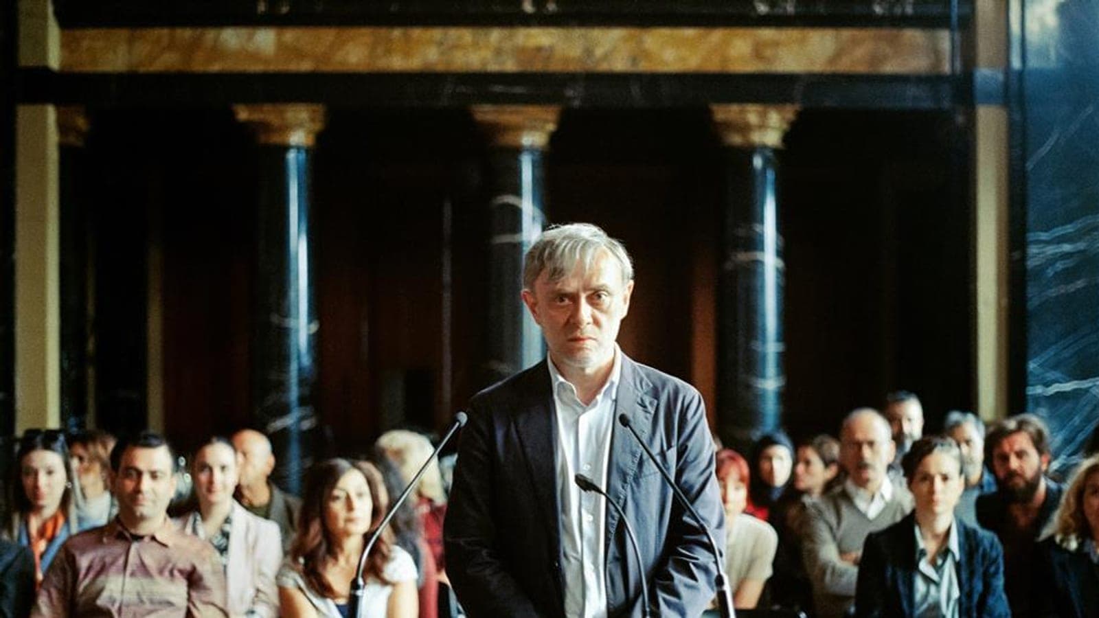 A grey-haired man in a dark jacket stands at a microphone facing forward, with a seated audience behind him in a panelled hall, in a still from Yellow Letters.