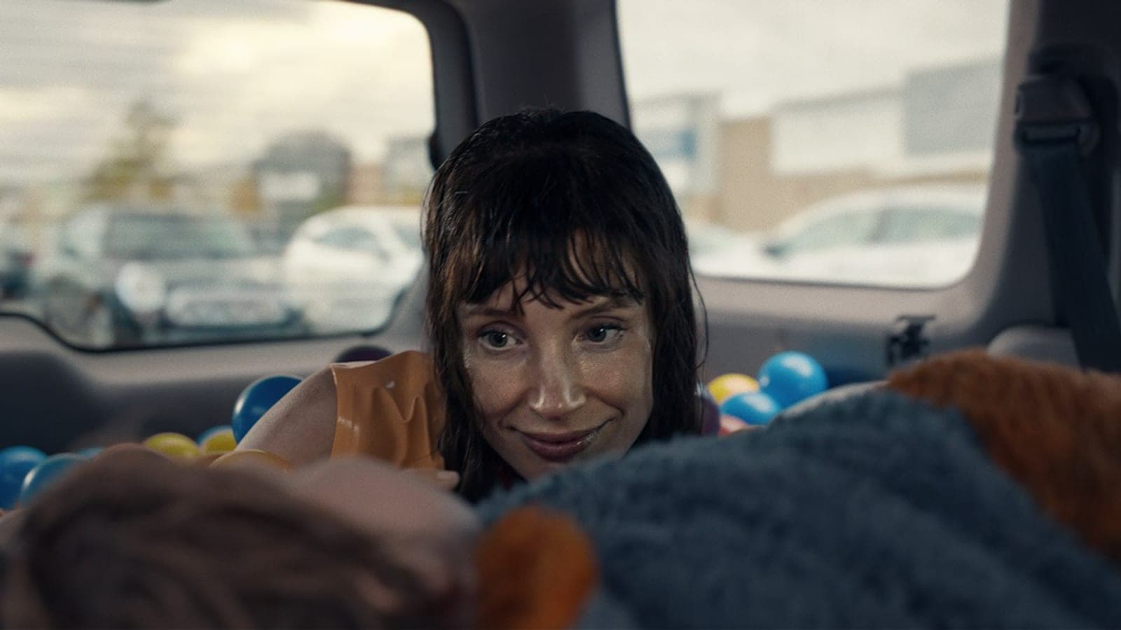 A woman with wet dark hair smiles over the back seat of a car strewn with coloured plastic balls, in a still from Other Mommy.