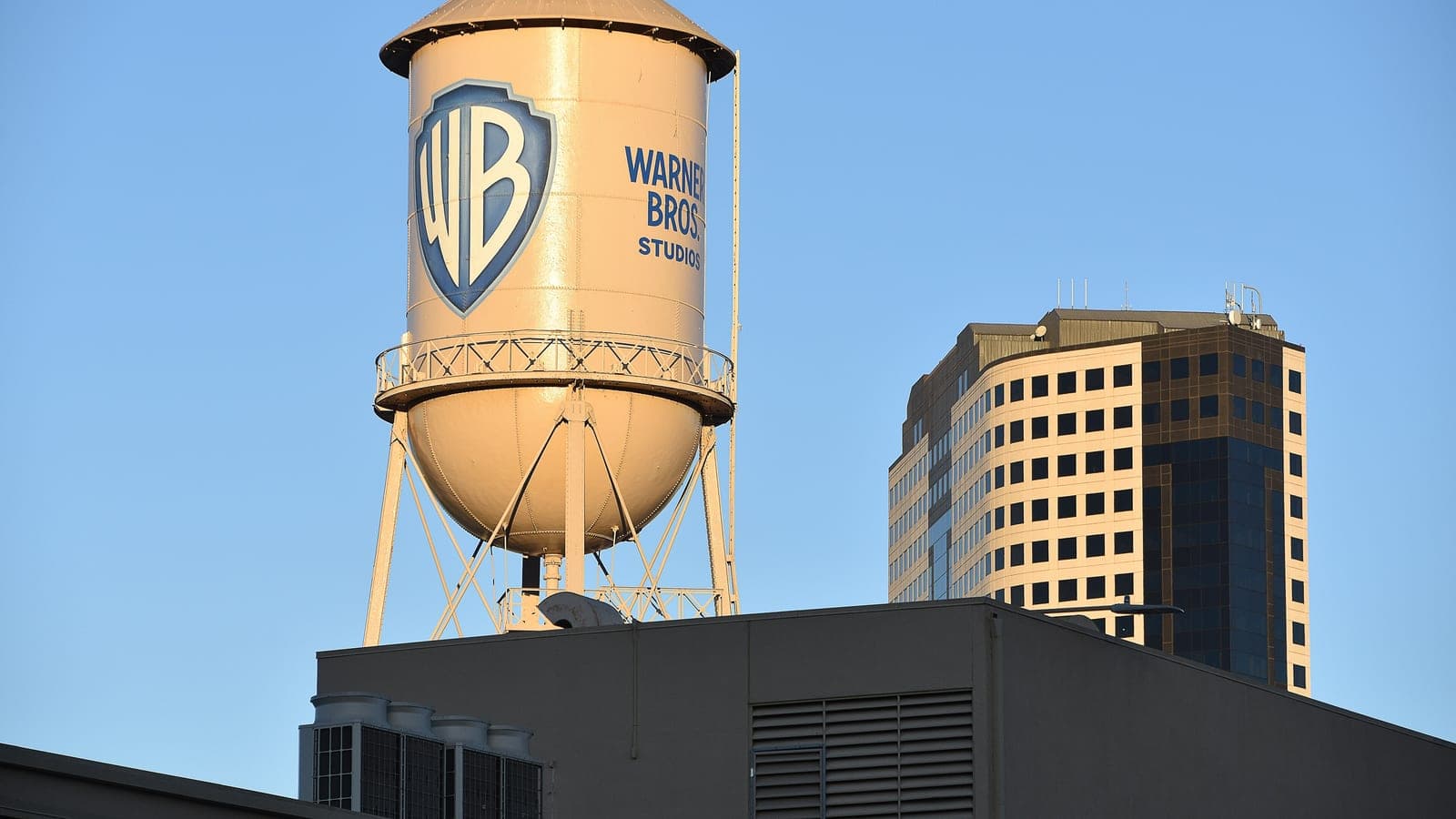The Warner Bros. Studios water tower in Burbank, California, lit by early sun against a clear sky, with an office block behind it.