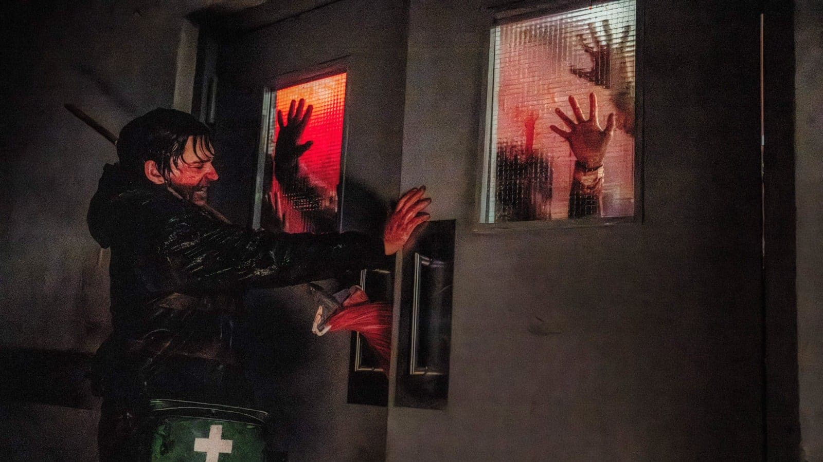 A blood-spattered man with a first-aid bag holds a door shut as hands press against the frosted windows behind him, in a still from Zach Cregger's Resident Evil