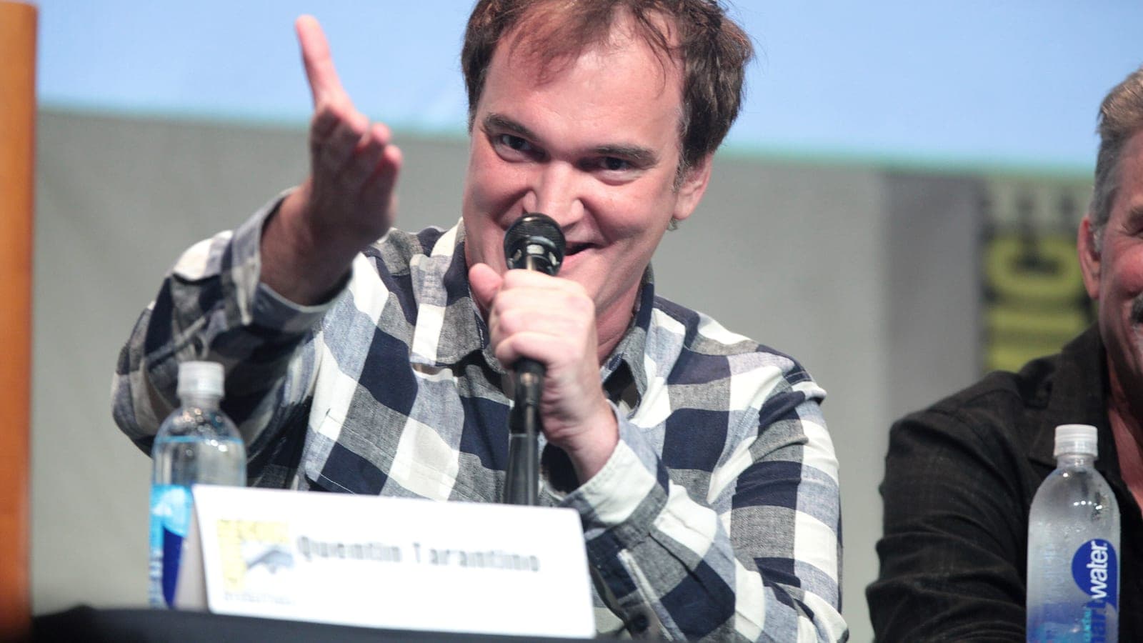 Quentin Tarantino in a checked shirt, gesturing with one hand while speaking into a microphone on a panel at San Diego Comic-Con in 2015