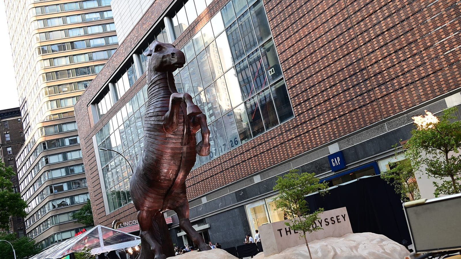 A towering horse sculpture outside the New York premiere of The Odyssey, the film title carved into a stone plinth beneath it.