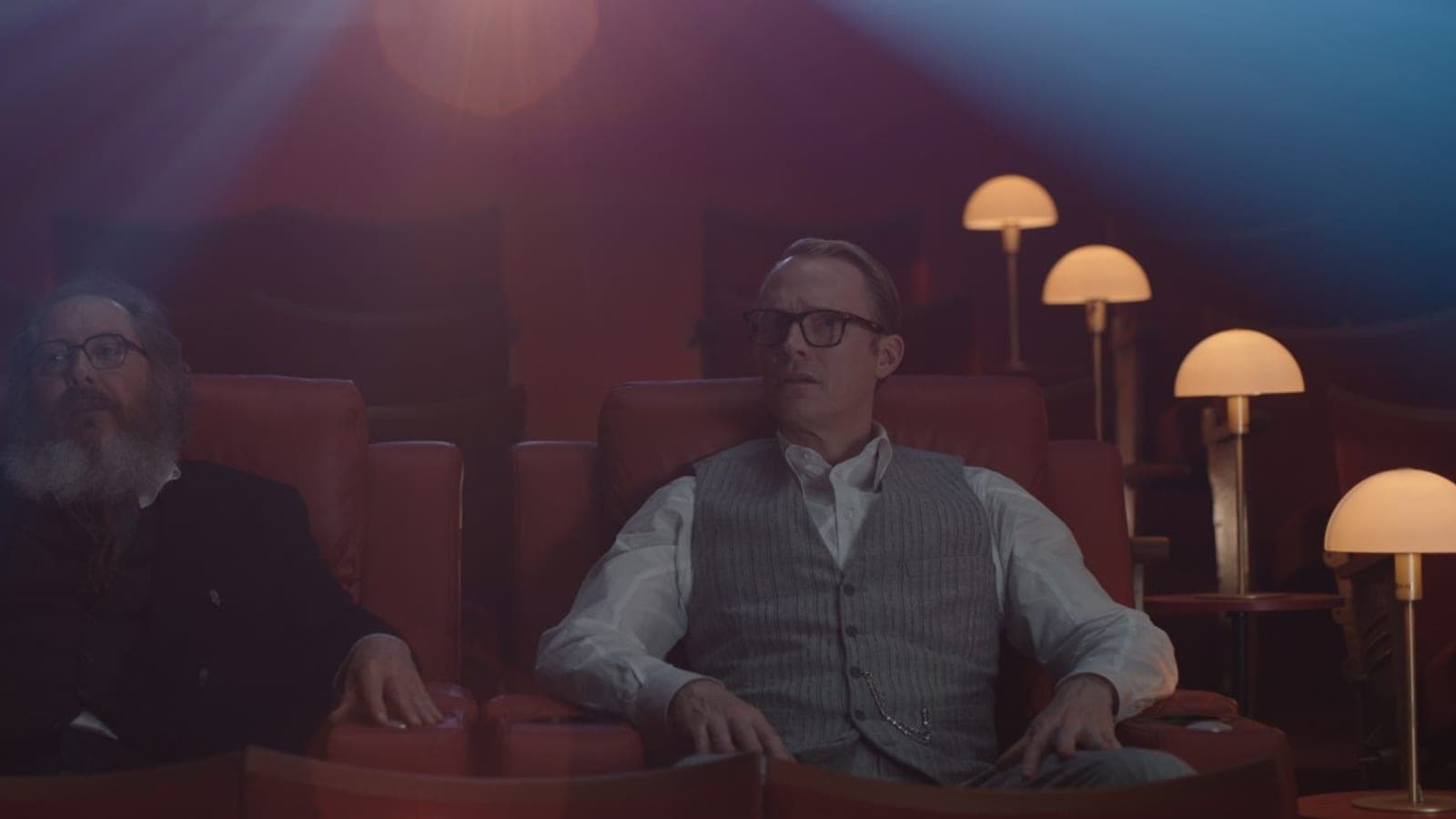 James Spader and Paul Bettany sit side by side in red cinema seats, lit by a projector beam, in a still from VisionQuest