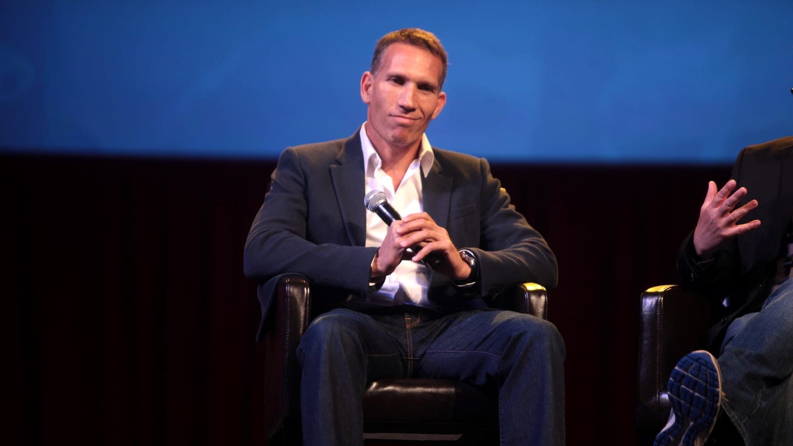 Ynon Kreiz sits in a leather armchair on a darkened panel stage, holding a microphone in both hands, in a dark jacket over an open-necked white shirt and jeans.